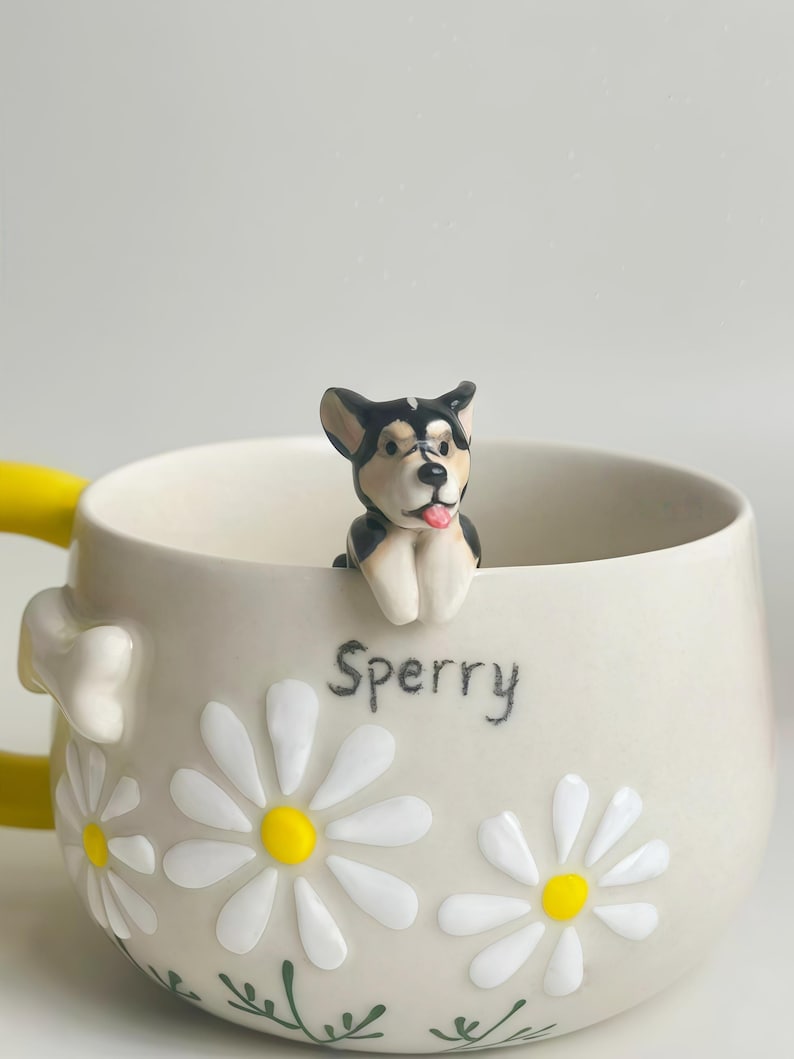 Puede incluir: Una taza de cer&aacute;mica color crema con una figura tridimensional de un perro en blanco y negro asom&aacute;ndose por el borde. La taza est&aacute; decorada con p&eacute;talos de margarita blancos en relieve con centros amarillos y el nombre Sperry en negro.