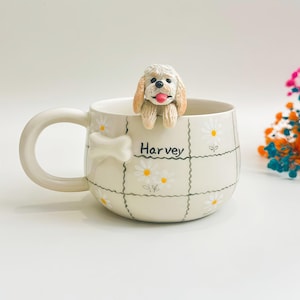 Könnte beinhalten: Ein cremefarbener Keramikbecher mit einem Gittermuster und weißen Gänseblümchen. Eine beige Hundefigur mit rosa Zunge ruht auf dem Rand. Der Name Harvey ist in schwarzer Schrift auf der Vorderseite aufgedruckt, ergänzt durch einen weißen Knochen.