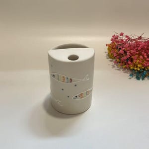 Puede incluir: Un jarrón cilíndrico de cerámica blanquecina con una parte superior curvada y un solo agujero. El jarrón está decorado con ilustraciones caprichosas y coloridas de peces. Un pequeño ramo de flores secas está en el fondo.