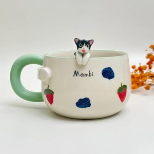 Op de afbeelding: Een crèmekleurige keramische mok met een lichtgroen handvat en een klein, gebeeldhouwd zwart-wit kattenfiguurtje op de rand. De mok is versierd met geschilderde aardbeien en blauwe bessen, met de naam Mambi in het zwart gedrukt.