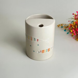May include: A cylindrical off-white ceramic vase featuring a flat top with a circular opening. The exterior is decorated with hand-painted fish motifs in orange, yellow, and blue, accompanied by small blue dots scattered across the surface.