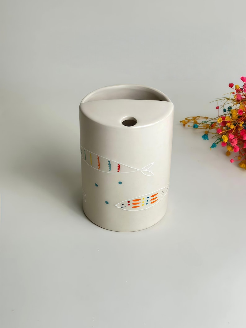 May include: A cylindrical off-white ceramic vase featuring a flat top with a circular opening. The exterior is decorated with hand-painted fish motifs in orange, yellow, and blue, accompanied by small blue dots scattered across the surface.