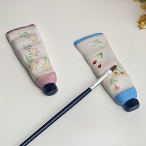 Puede incluir: Dos figuras de cerámica con forma de tubos de pintura con diseños florales y un pincel. Un tubo tiene una base rosa y una parte superior morada, el otro tiene una parte superior azul. El pincel tiene un mango azul oscuro y cerdas plateadas.