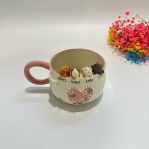 May include: A cream-colored ceramic mug with a pink handle and rim, decorated with a heart and floral design. The mug contains small animal figurines and the names "BERTI", "MIKE", and "LAIA".