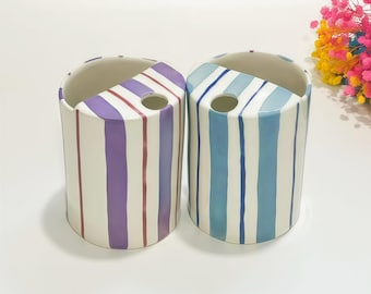 May include: Two cylindrical ceramic toothbrush holders with a semi-circular top opening and a small circular hole. One holder features vertical purple and maroon stripes on a white base, while the other has vertical blue and light blue stripes.