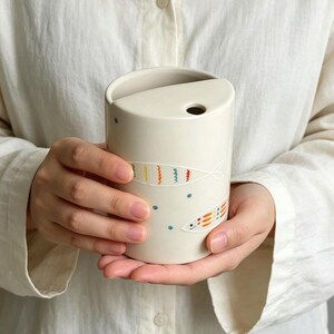 May include: A cylindrical off-white ceramic cup featuring a unique semi-circular lid with a small circular opening. The exterior is decorated with hand-painted colorful fish motifs in orange, yellow, and blue, accented by small blue dots.