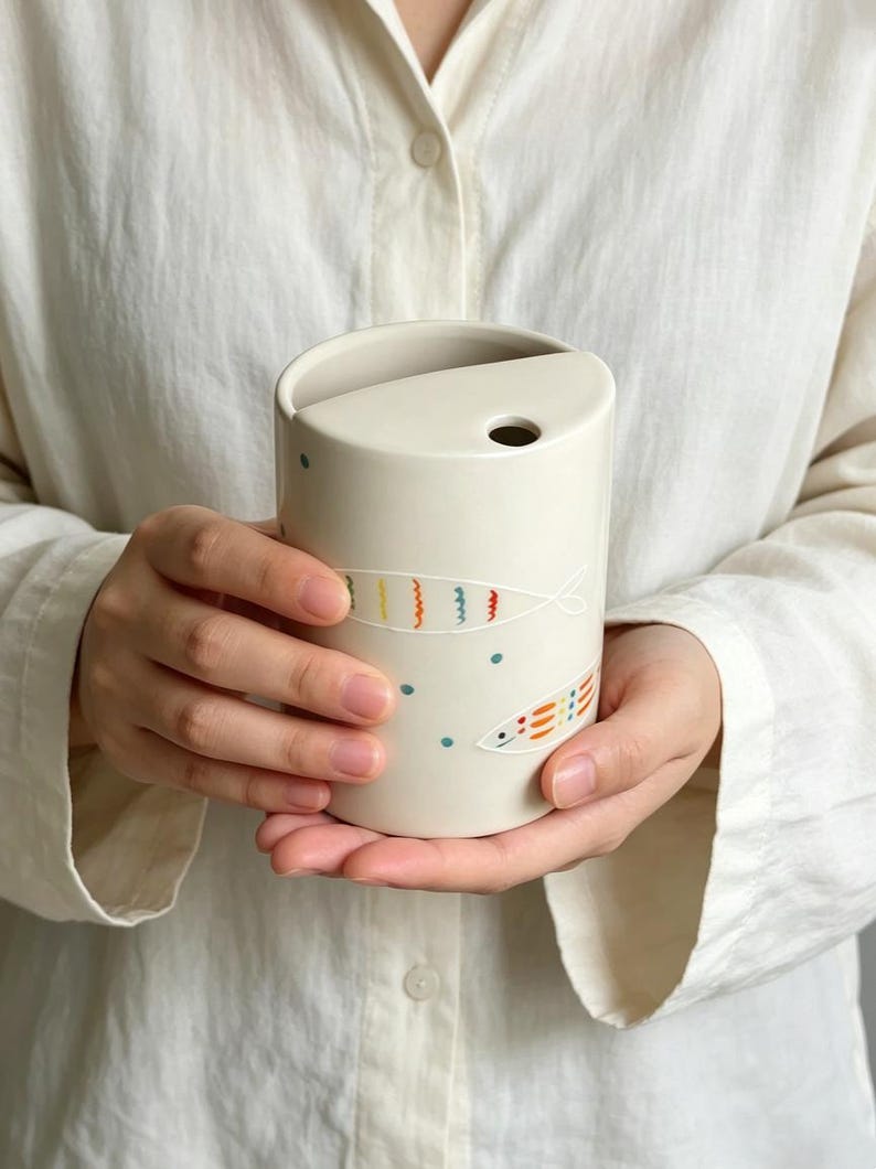 May include: A cylindrical off-white ceramic cup featuring a unique semi-circular lid with a small circular opening. The exterior is decorated with hand-painted colorful fish motifs in orange, yellow, and blue, accented by small blue dots.