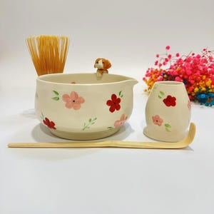 Könnte beinhalten: Ein Matcha-Teeset mit einer cremefarbenen Schale und einem passenden Halter, beide mit rosa Blumenmustern verziert. Eine kleine Hundefigur sitzt am Rand der Schale. Enthält einen Bambusbesen und -löffel.