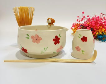 May include: A ceramic matcha bowl with a small brown and white dog figurine on the rim, decorated with pink and red flowers and green leaves. Includes a bamboo whisk, a bamboo scoop, and a matching ceramic whisk holder, all featuring floral motifs.