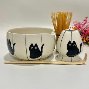 May include: A cream-colored ceramic matcha bowl, whisk holder, and scoop set. The bowl and holder feature black cat illustrations and vertical black lines. A bamboo whisk and scoop are also included. The set is designed for preparing matcha tea.