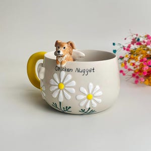 Puede incluir: Una taza de cer&aacute;mica beige con una figura esculpida de un perro marr&oacute;n y blanco en el borde, un asa amarilla brillante y margaritas blancas con centros amarillos pintadas en el lateral. El texto Chicken Nugget est&aacute; impreso en negro al frente.