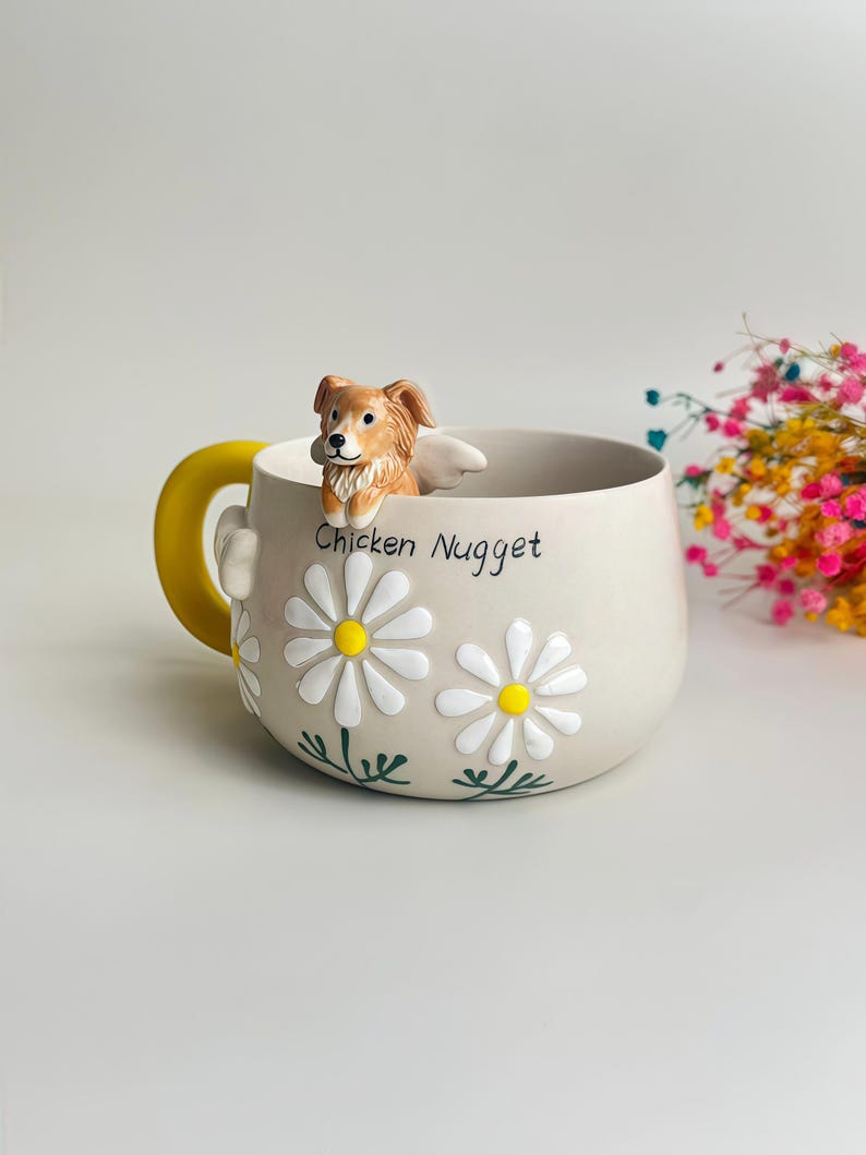 Puede incluir: Una taza de cer&aacute;mica beige con una figura esculpida de un perro marr&oacute;n y blanco en el borde, un asa amarilla brillante y margaritas blancas con centros amarillos pintadas en el lateral. El texto Chicken Nugget est&aacute; impreso en negro al frente.