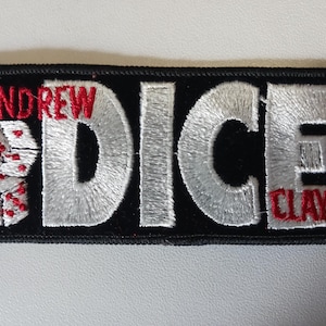 Könnte beinhalten: Gestickter Aufnäher mit den Worten "ANDREW DICE CLAY" in rotem und weißem Faden. Das Wort "DICE" ist in großem, silbernem Faden gehalten. Zwei Würfel mit roten Punkten befinden sich auf der linken Seite. Der Aufnäher ist rechteckig mit schwarzem Hintergrund.