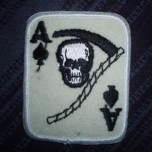 Puede incluir: Un parche de fieltro blanco con una imagen bordada en negro del As de Picas. La carta presenta una calavera con una guadaña y la letra 'A' en la esquina.