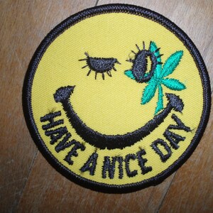 May include: Embroidered yellow patch with a smiling face design. The patch features a black smiling mouth, a winking eye, and a green cannabis leaf with a black sunflower. The words "HAVE A NICE DAY" are embroidered in black.