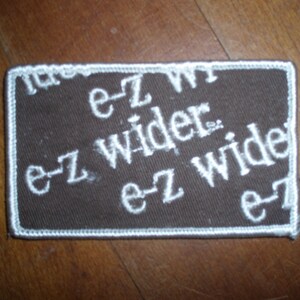 May include: Brown fabric patch with white embroidered text that reads "e-z wider e-z wide e-z".