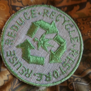 May include: A white circular patch with green embroidered text and a green embroidered recycling symbol. The text reads "Reduce - Reuse - Recycle".