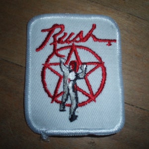May include: White embroidered patch with the word "Rush" in red cursive lettering and a red star with a figure in white standing in the center.