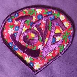 Peut inclure: Écusson en forme de cœur avec le mot "LOVE" brodé en fil violet. Le fond présente un motif floral avec des fleurs rouges, blanches, bleues et jaunes. L'écusson est bordé d'une bordure violette.