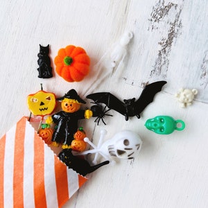 May include: A collection of small Halloween-themed plastic charms and figures spilling from an orange and white striped paper bag. The items include a black cat, orange pumpkin, scarecrow, bat, spider, green skull, white skull, and ghost figures.