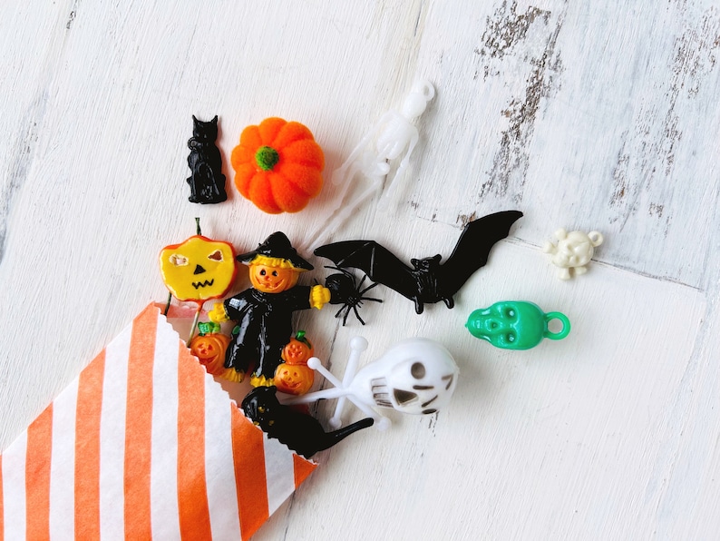 May include: A collection of small Halloween-themed plastic charms and figures spilling from an orange and white striped paper bag. The items include a black cat, orange pumpkin, scarecrow, bat, spider, green skull, white skull, and ghost figures.