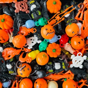 May include: A collection of small plastic Halloween-themed charms and figurines in orange, black, white, green, and blue. The assortment includes miniature pumpkins, skulls, bats, owls, ghosts, and various body parts scattered in a dense pile.