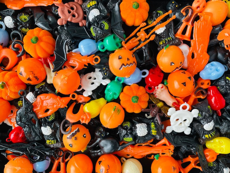 May include: A collection of small plastic Halloween-themed charms and figurines in orange, black, white, green, and blue. The assortment includes miniature pumpkins, skulls, bats, owls, ghosts, and various body parts scattered in a dense pile.