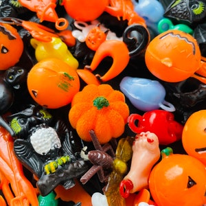 May include: A collection of assorted plastic Halloween-themed charms and figurines including orange pumpkins, black cats, skulls, a fuzzy orange pumpkin, a black owl, a severed foot, and various bats in bright orange, black, white, and blue colours.