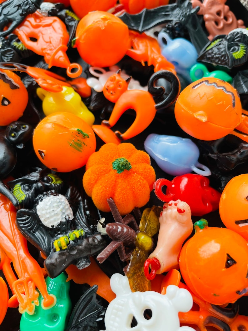 May include: A collection of assorted plastic Halloween-themed charms and figurines including orange pumpkins, black cats, skulls, a fuzzy orange pumpkin, a black owl, a severed foot, and various bats in bright orange, black, white, and blue colours.