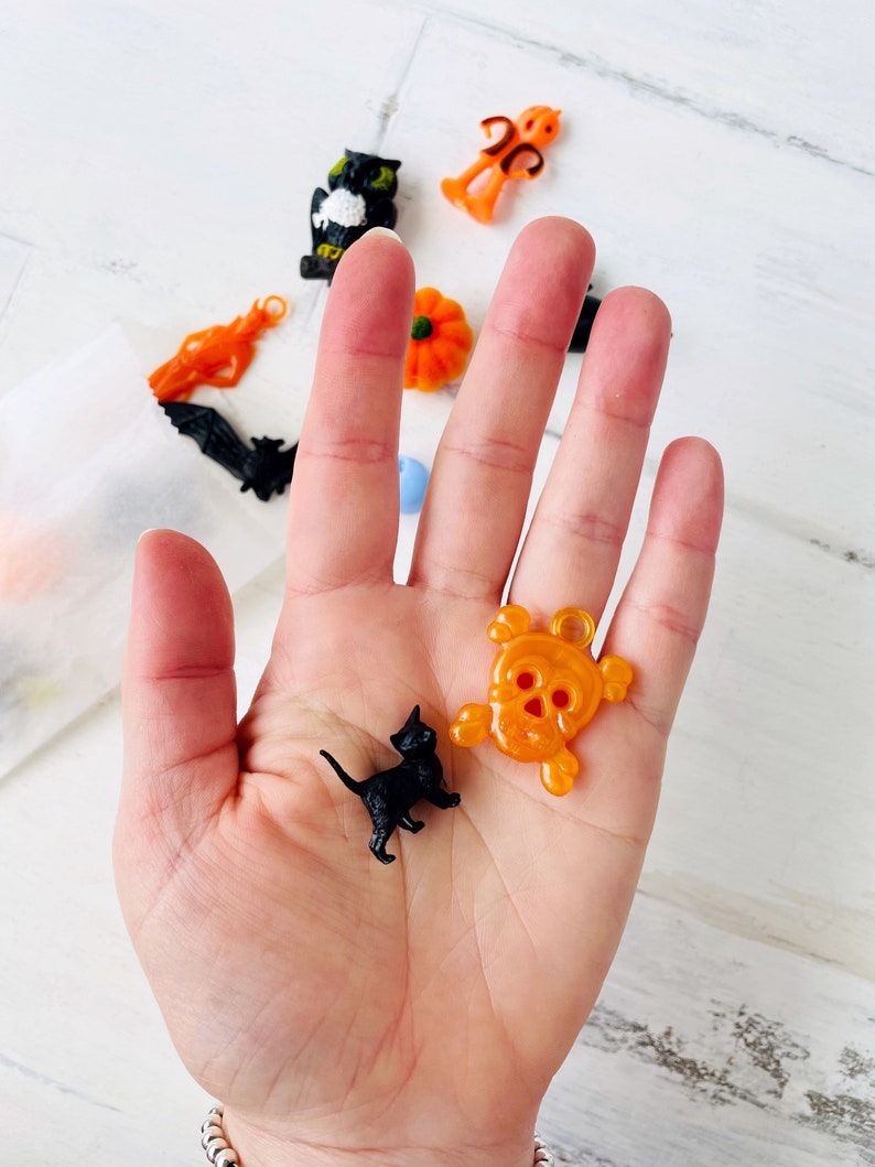 May include: A small black cat figurine and an orange translucent skull-shaped charm. The cat is in a walking pose with its tail raised. The skull charm features a loop at the top for hanging. Additional small Halloween-themed charms are visible.