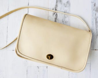 May include: A cream-colored leather shoulder bag with a gold-toned clasp. The bag has a simple design and a long strap.