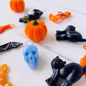 May include: A collection of small Halloween-themed charms including orange pumpkins, black cats, a blue skull, a black bat, and orange figures. Each charm features a small loop at the top for attachment, showcasing various festive shapes and colours.