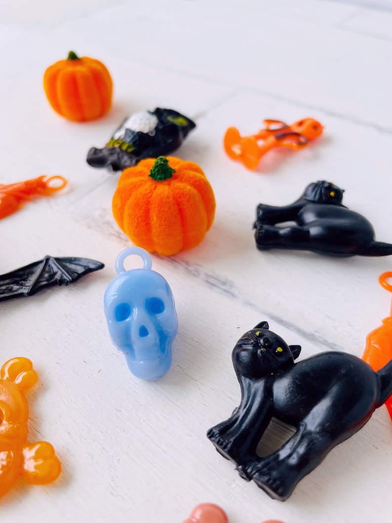 May include: A collection of small Halloween-themed charms including orange pumpkins, black cats, a blue skull, a black bat, and orange figures. Each charm features a small loop at the top for attachment, showcasing various festive shapes and colours.