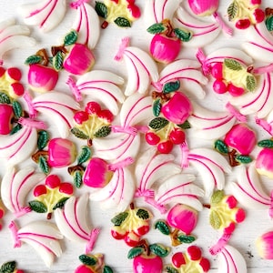 May include: A collection of white and pink fruit-shaped embellishments, including bananas, apples, and cherries. The bananas have pink stripes, and the apples and cherries are bright pink. Green and yellow leaves add detail to the fruit.