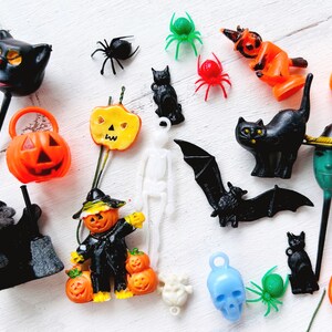 May include: A collection of miniature Halloween-themed plastic figurines and charms, including black cats, orange pumpkins, spiders, a white skeleton, a black bat, a blue skull, and green spiders, arranged in a scattered assortment on a white surface.