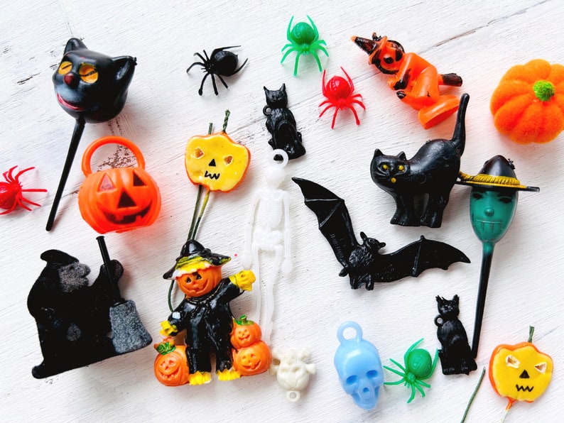 May include: A collection of miniature Halloween-themed plastic figurines and charms, including black cats, orange pumpkins, spiders, a white skeleton, a black bat, a blue skull, and green spiders, arranged in a scattered assortment on a white surface.