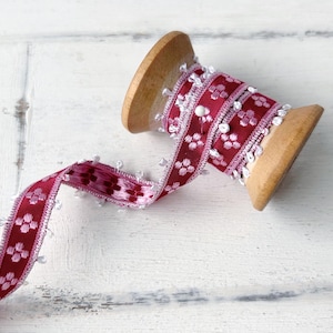 Pode incluir: Um carretel de fita vermelha e branca com um padrão floral e contas brancas ao longo da borda. A fita está enrolada em torno de um carretel de madeira.