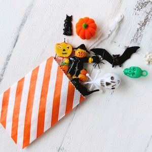 May include: An orange and white striped paper bag spilling out a collection of small Halloween-themed plastic charms, including a black cat, an orange pumpkin, a scarecrow, a black bat, a green skull, a white skull, and a white ghost figure.