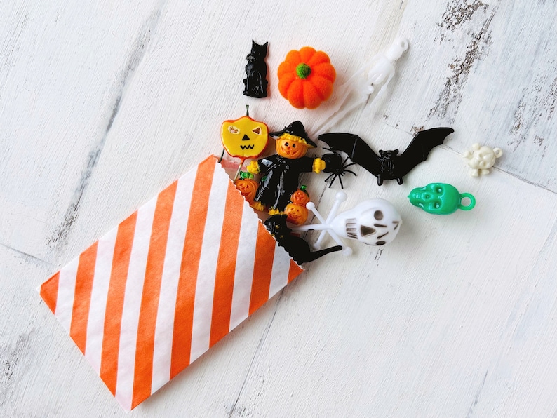 May include: An orange and white striped paper bag spilling out a collection of small Halloween-themed plastic charms, including a black cat, an orange pumpkin, a scarecrow, a black bat, a green skull, a white skull, and a white ghost figure.