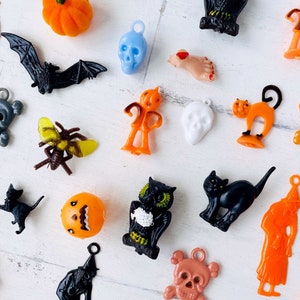 May include: A collection of assorted plastic Halloween charms and pendants featuring various figures including orange pumpkins, black bats, skulls in blue, white, and copper, a black cat, an owl, a witch, a spider, and a severed foot with red nail polish.