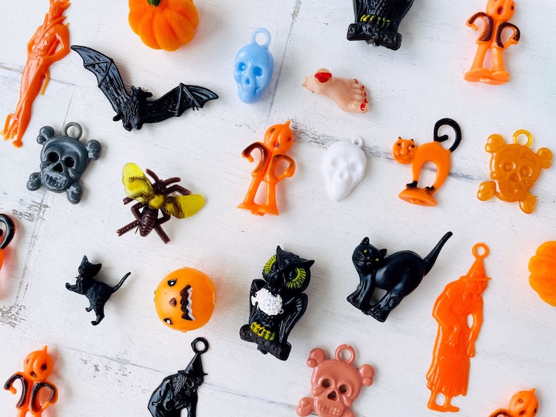 May include: A collection of assorted plastic Halloween charms and pendants featuring various figures including orange pumpkins, black bats, skulls in blue, white, and copper, a black cat, an owl, a witch, a spider, and a severed foot with red nail polish.