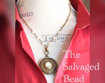 May include: A gold necklace with a large, round pendant featuring a white circle in the center. The pendant has a decorative, swirling design. The necklace is on a white mannequin wearing a red shirt. The text "The Salvaged Bead" is visible in the image.