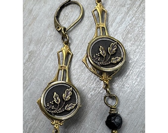 May include: A pair of gold tone earrings with black gemstone accents. Each earring features a round, black cameo with a gold floral design. The earrings are dangle style with a black gemstone drop.