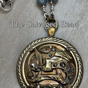 May include: A gold-toned pendant necklace with a round, ornate, metal charm featuring a stylized letter 'L' with a rope-like border. The charm is decorated with small, black rhinestones. The necklace is made with a silver chain and blue glass beads.