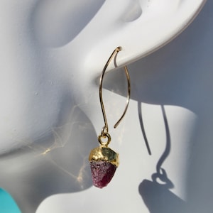 May include: Gold earrings with a pink gemstone pendant. The earrings are a simple design with a small, delicate hook.