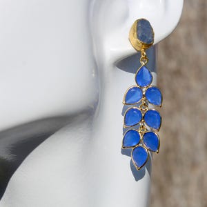 May include: Gold-toned dangle earrings featuring a blue stone at the top and a cascade of blue teardrop-shaped stones. The earrings are displayed against a white background, showcasing their design and color.