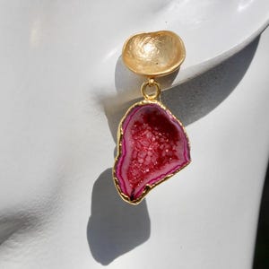 May include: A single gold-colored earring with a pink and red geode stone pendant. The geode has a gold-colored trim and is suspended from a gold-colored, shell-shaped stud.