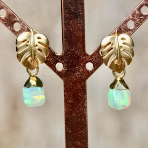 May include: A pair of gold-toned earrings featuring a monstera leaf stud design with a dangling, raw-cut, iridescent white opal-like stone. The earrings are displayed on a copper-colored metal stand, showcasing the metallic finish and stone texture.