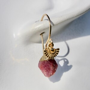 May include: A gold-colored earring with a raw, faceted, deep pink gemstone. The earring has a small, decorative gold element above the gemstone. The earring is shown on a white background.