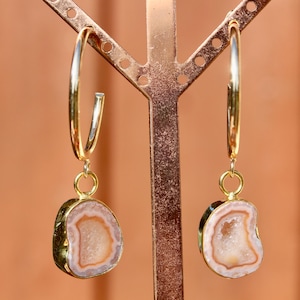 May include: A pair of gold hoop earrings featuring dangling, irregular-shaped agate slices. Each slice displays concentric bands of soft pink and white tones with a textured, crystalline center, suspended from a polished gold-tone metal hoop.